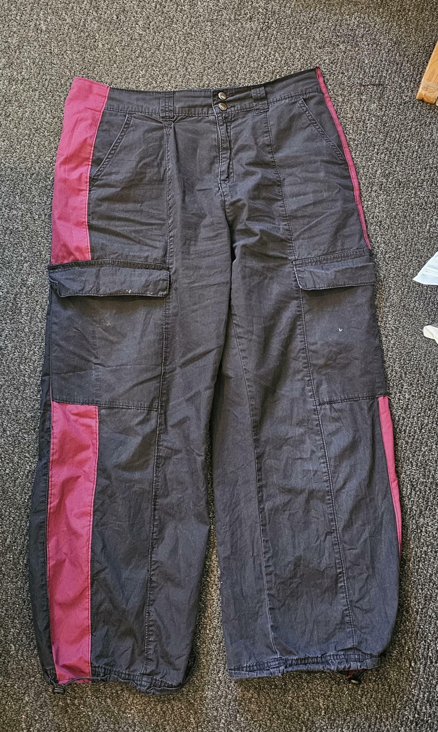 A pair of black cargo pants laid out on the floor. There is a stripe of red fabric running down each side of the pant legs.
