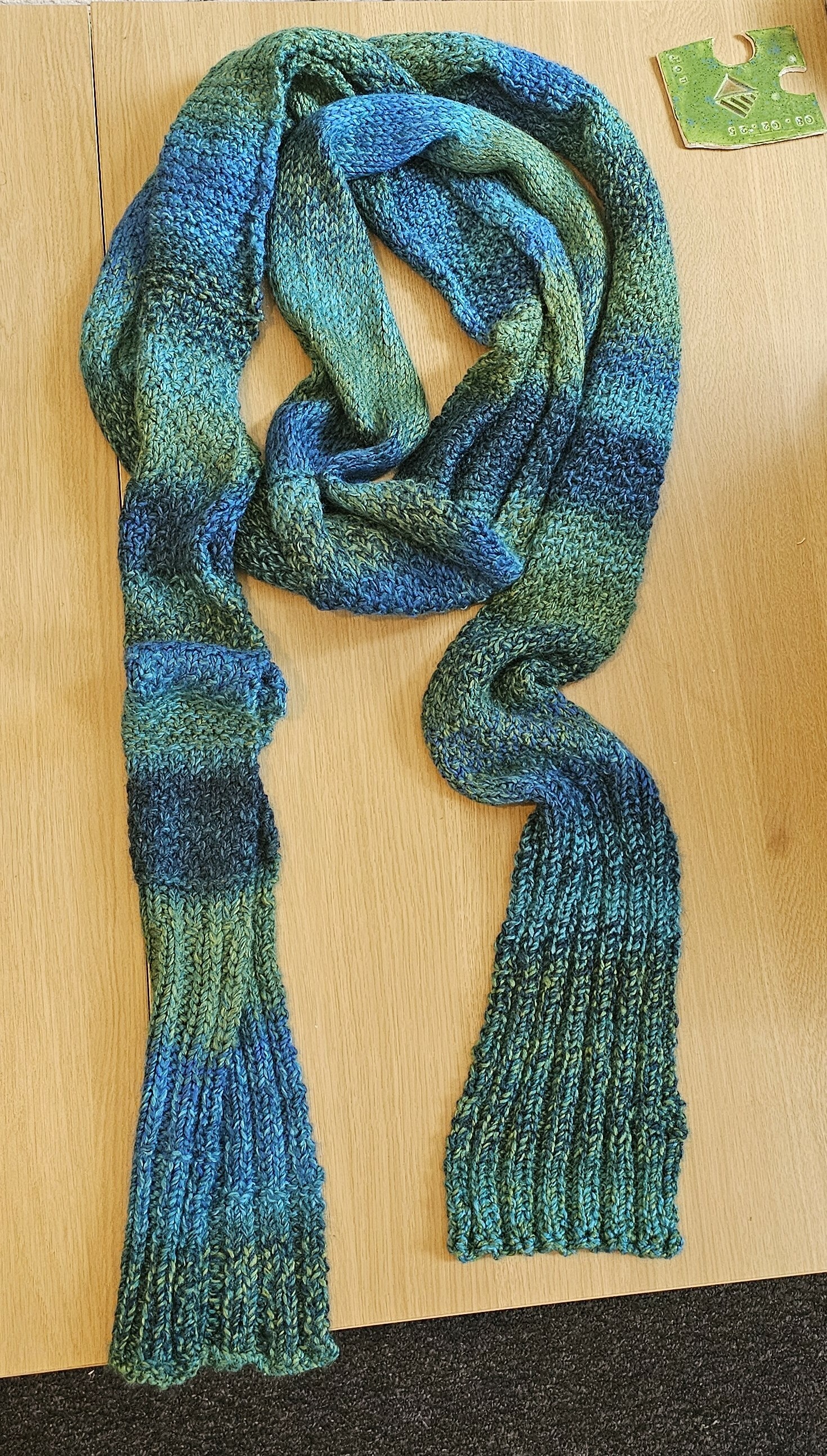 A top-down photo of a long scarf laid out on a table. It is a gradient of colours ranging from blue, to teal, to green.