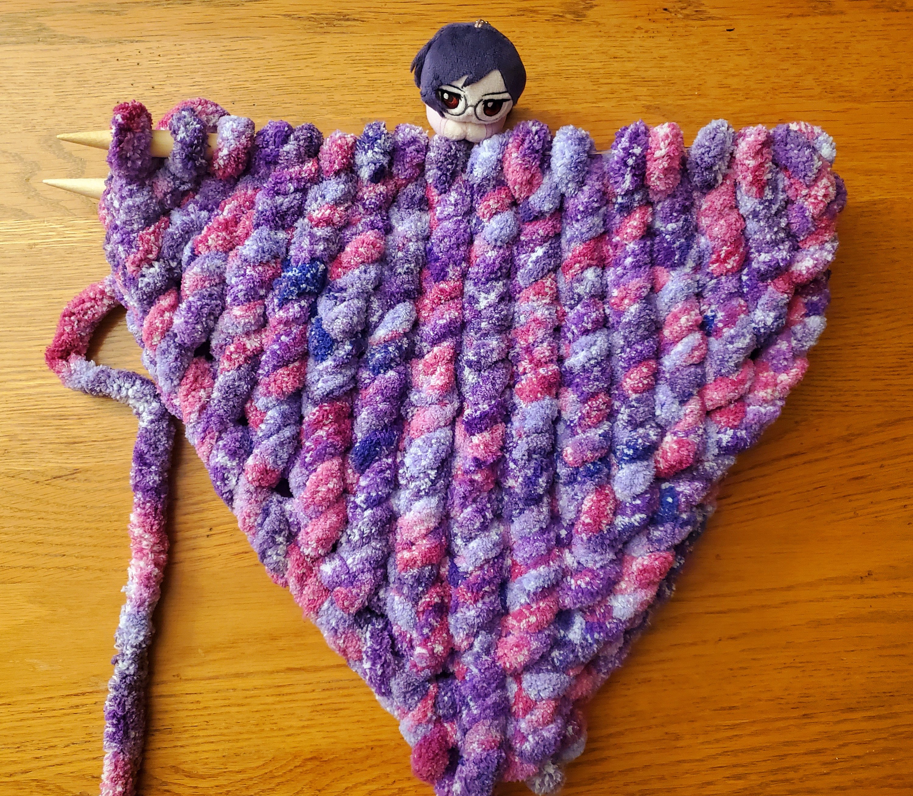 A half finished pillow made of jumbo yarn that is purple. A Tieria plush sits next to the working row.