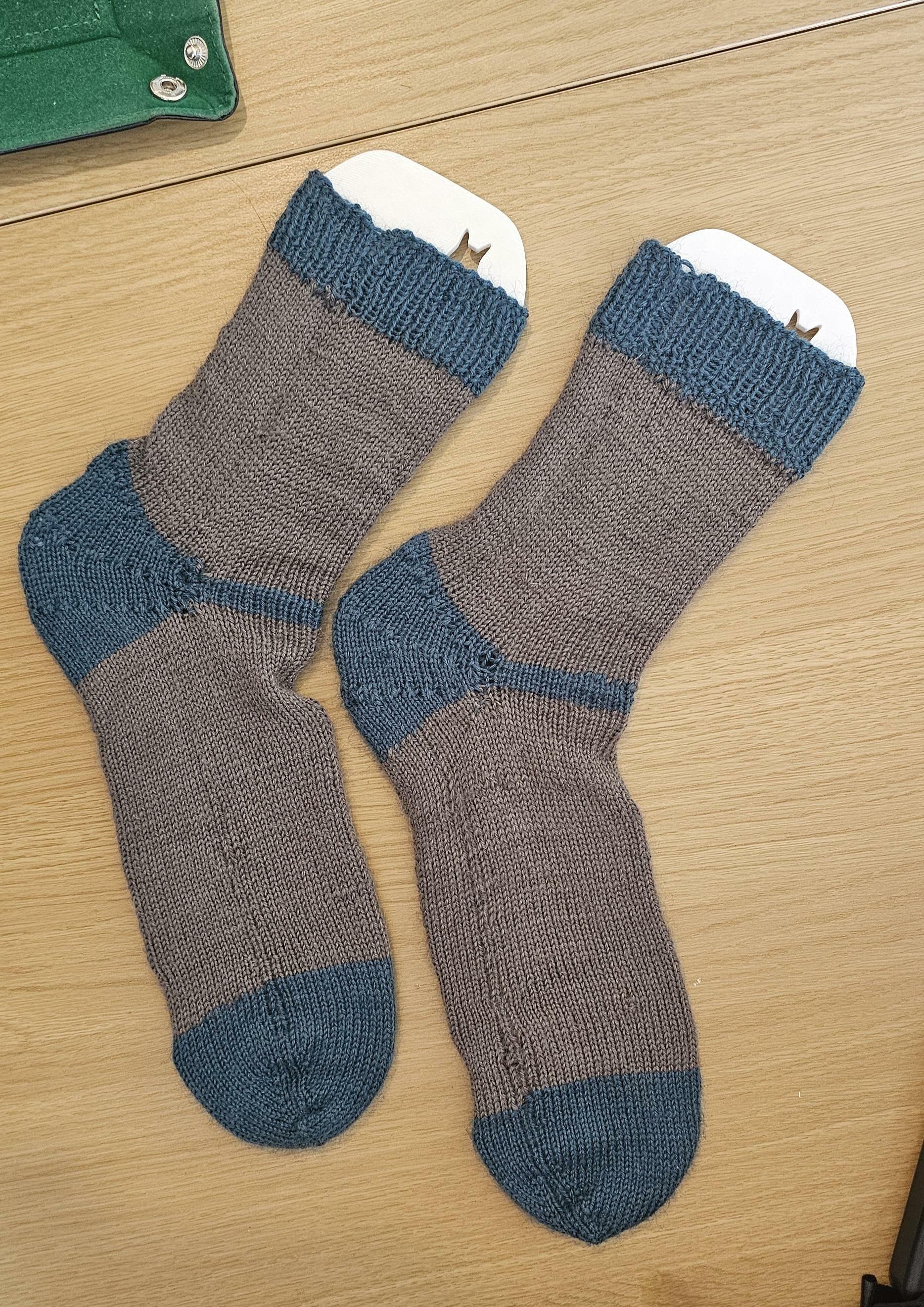 A pair of grey socks sitting side by side in a sock block. The body of the socks are grey and the toe, heel, and cuff are a teal blue colour.