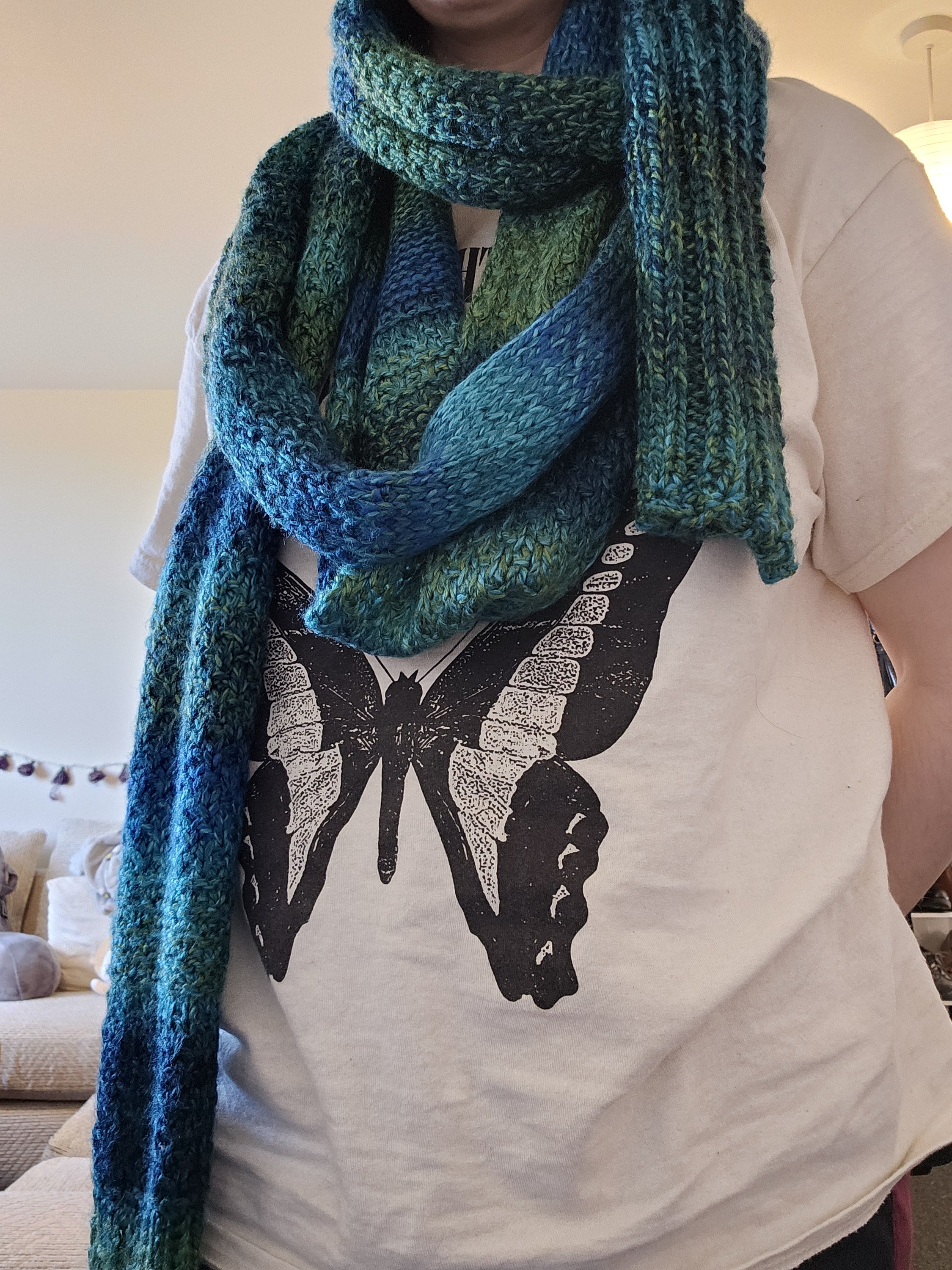 A front facing picture of a person with a very long scarf wrapped many times around them. It is a gradient of colours ranging from blue, to teal, to green.